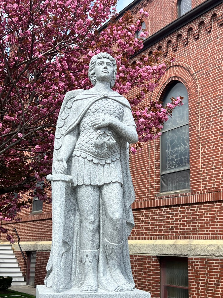Saint Michael the Archangel statue in front of church with tree in bloom with pink flowers.