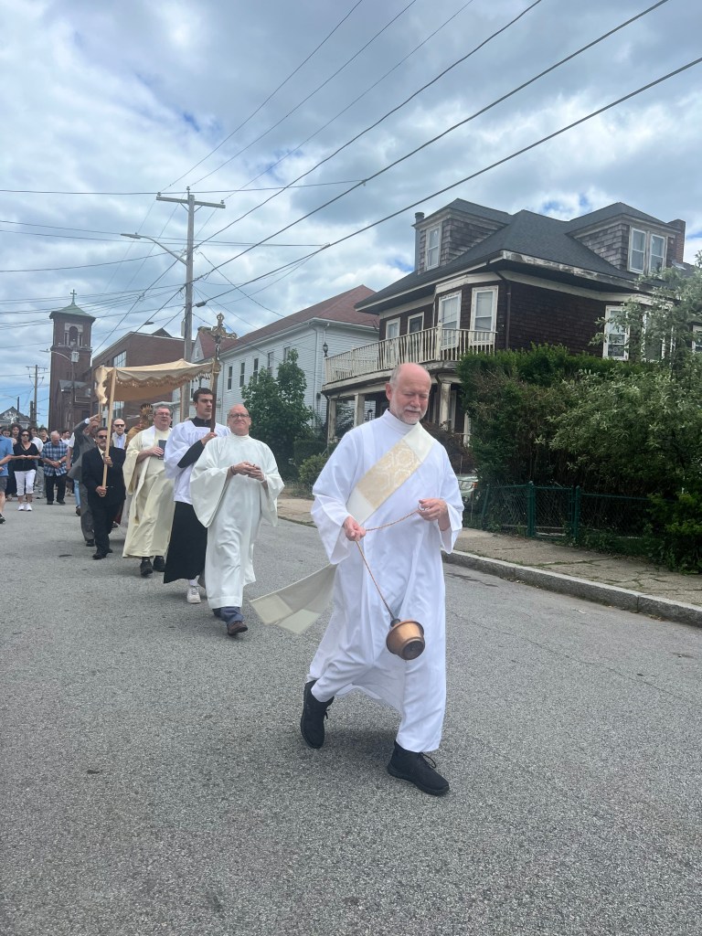 A Catholic deacon walking on a street holding a censer followed by a priest and others holding the Blessed Sacrament