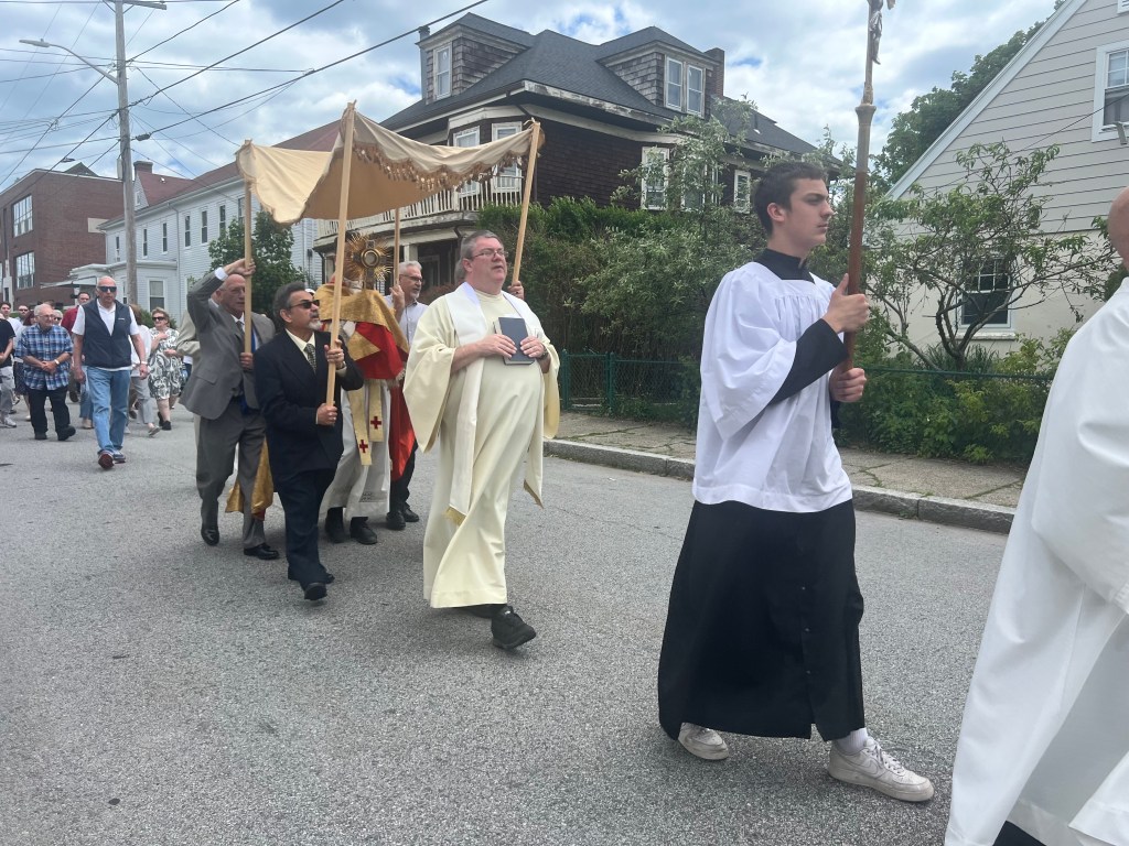 Outdoor procession lead by an altar boy carrying a crucifix, followed by a priest, followed by canopy bearers under which another priest holds the Blessed Sacrament.